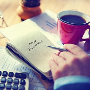 A businessman focusedly writes on a notebook the word New Business with a pen, accompanied by a cup of coffee nearby.